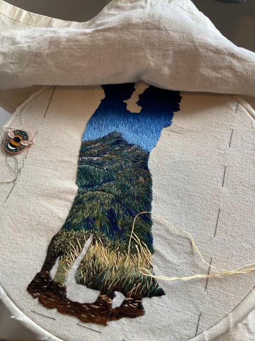 Embroidery in progress on a hoop. The embroidery depicts the full body silhouettes of a couple. The filling is a landscape with mountains.