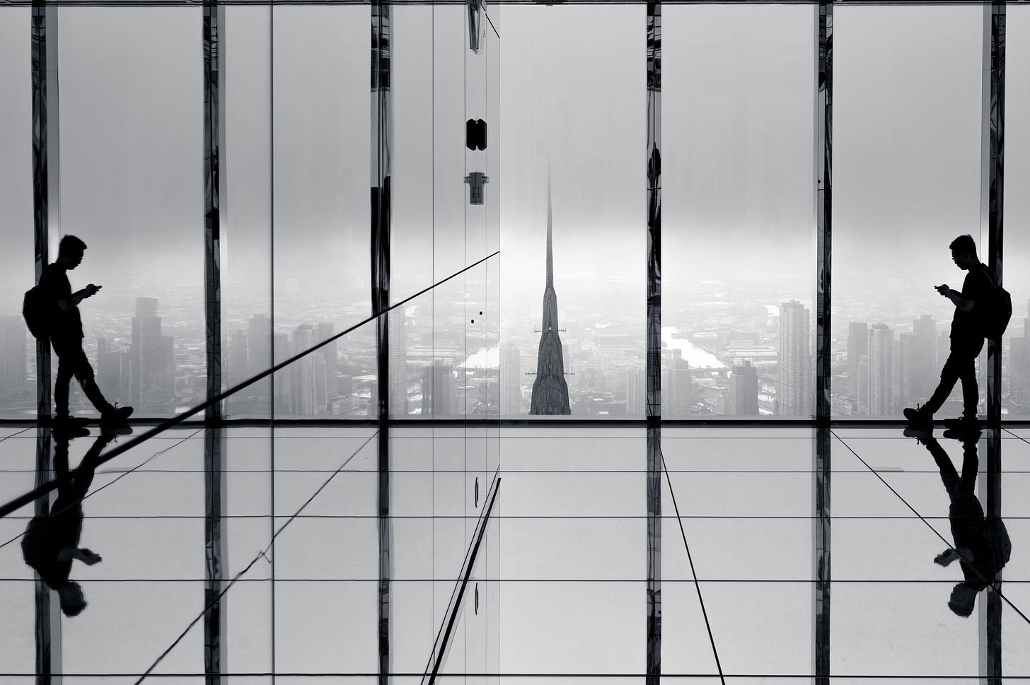 A man on his phone silhouetted against floor to ceiling glass and reflected symmetrically on both sides of the mirrored room at Summit One Vanderbilt, the Chrysler Building spire in fog beyond, New York, in black and white.
