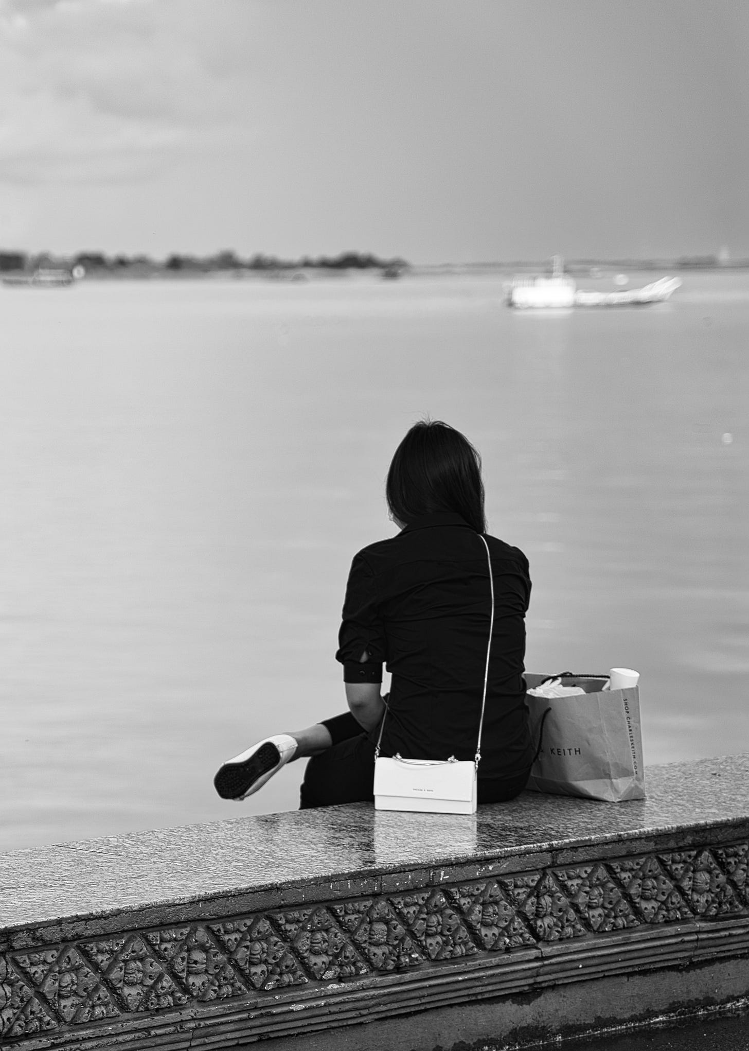 B&W picture of a woman sitting with her back to the camera, looking at a large body of water, possibly a lake.