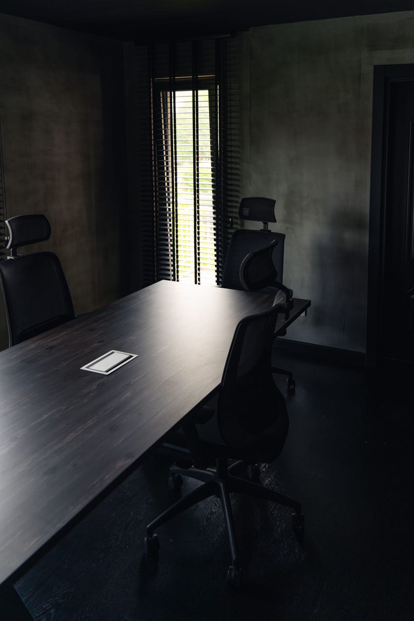 A dark conference room. The image frame is vertical and narrow, and a narrow window echoes the shape in the background while also seeming to be the only light source. Three black office chairs with headrests sit around a long, dark table, which is empty except for a small white paper with a black rectangle around the edge. 
