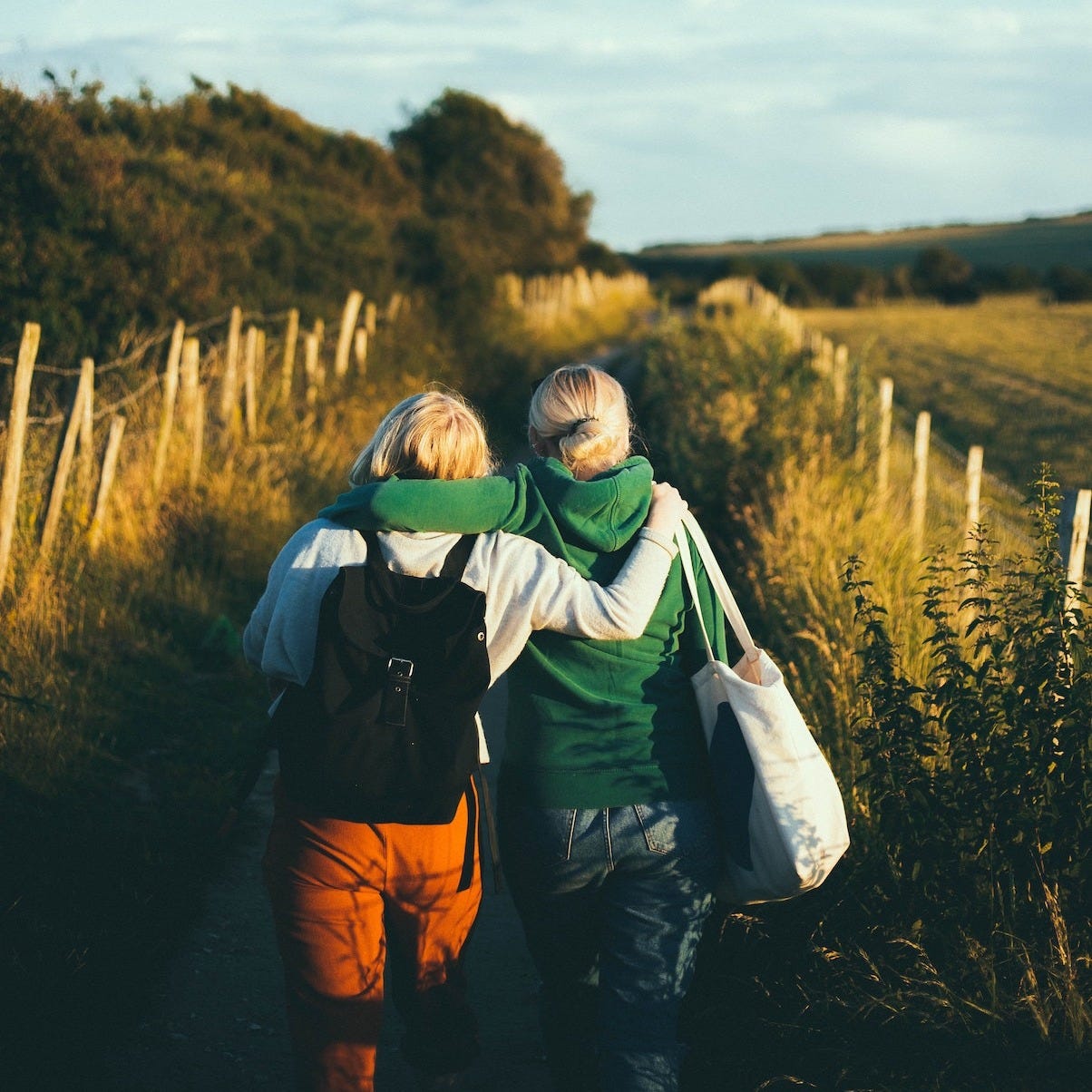 two woman walking on a trail by a field with their arms around each other two woman walking on a trail by a field with their arms around each other