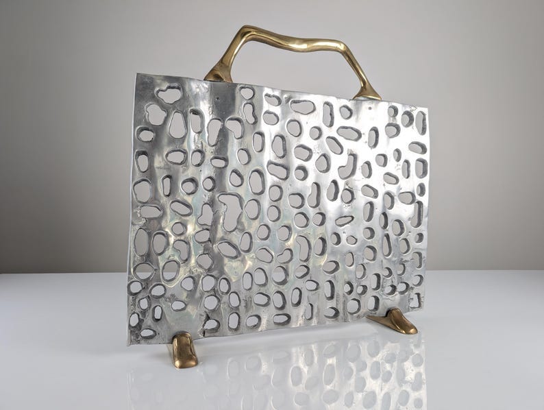 May include: A silver metal fireplace screen with a perforated design. The screen has a brass handle and two brass feet. The screen is standing upright on a white surface.