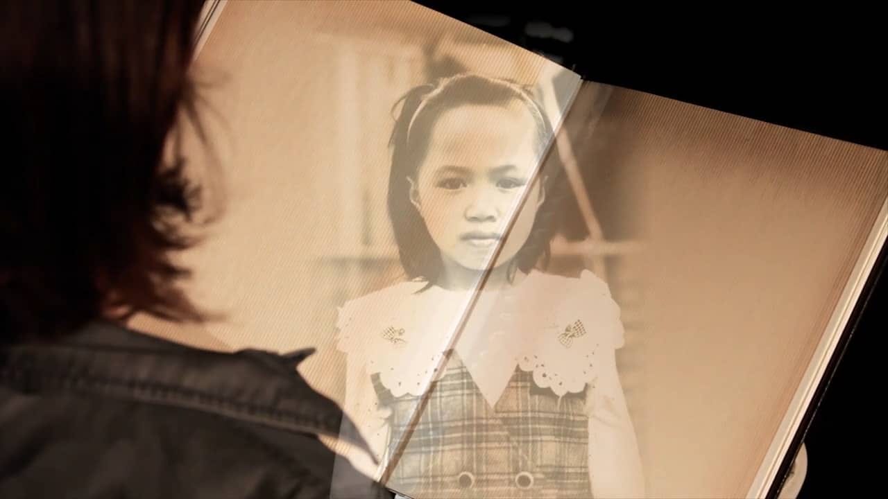 Een foto van een filmbeeld. Je kijkt over de schouders van een vrouw, die een groot boek vasthoud waarin een foto van een meisje van ongeveer 5 jaar is afgebeeld. Ze heeft een aziatisch uiterlijk en een gestreept jurkje aan en een band in haar haar.