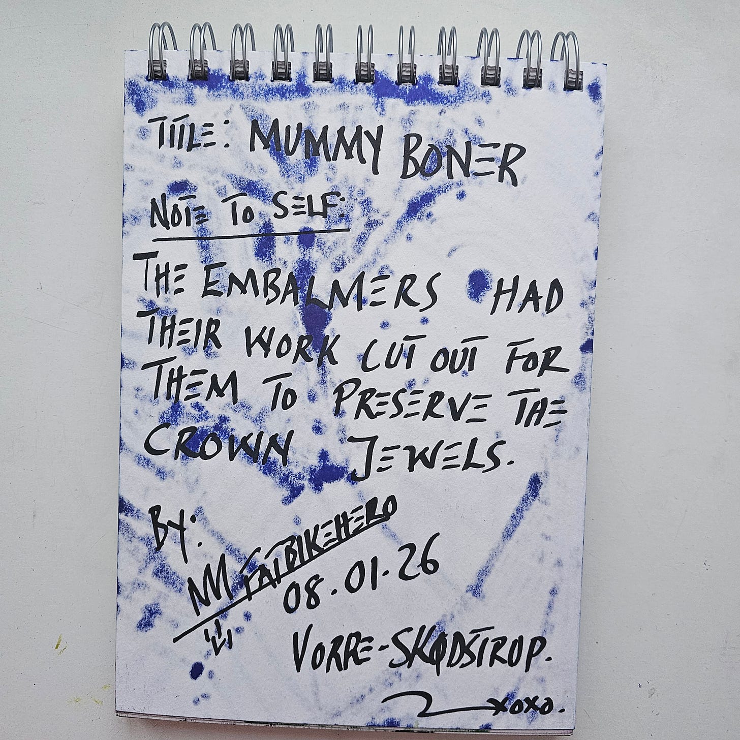 "The back of a sketchbook page featuring handwritten text in black marker. The text reads: 'Title: MUMMY BONER. Note to Self: The embalmers had their work cut out for them to preserve the crown jewels.' It is signed 'M FatbikeHero', dated '08.01.26', and inscribed 'Vorre - Skødstrup'. Blue ink from the front of the artwork has bled through the paper, creating a textured, spotted background."