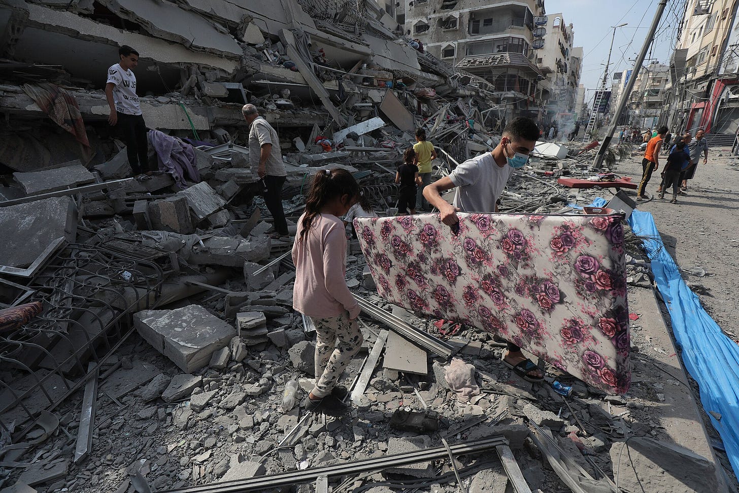 People stand amid the rubble of a building and looking at the ground. A man is carrying a large flower-patterned object.