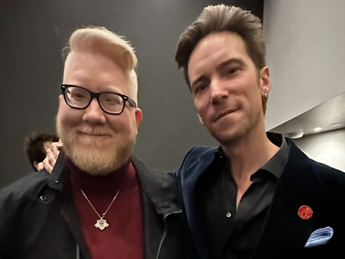 Photo of me and Troy Baker. I'm wearing a black zip up jacket with a red turtleneck and a silver pendant. Troy is wearing a black velvet suit jacket, black button up shirt that has a few top buttons unbuttoned and high waisted grey dress pants. He's holding a white bag with the Kojima Productions logo on it. 