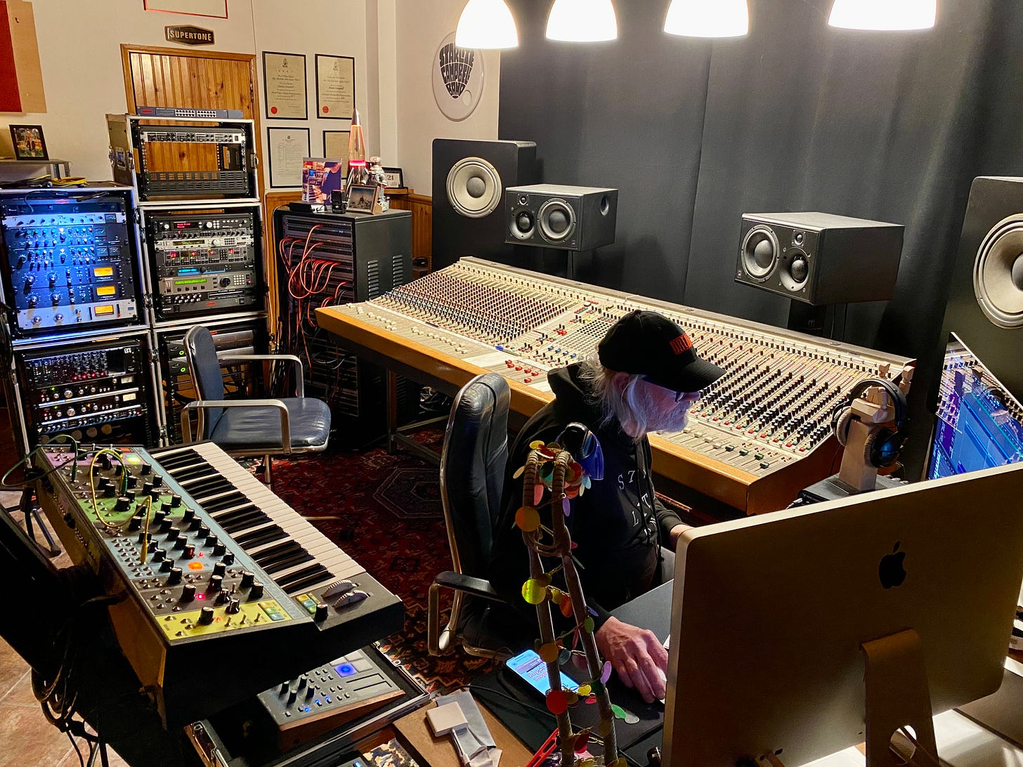 Man sat at a desk in a Supertone recording studio with a big CADAC console with faders and buttons. Keyboards on the left and electronic gear at the back