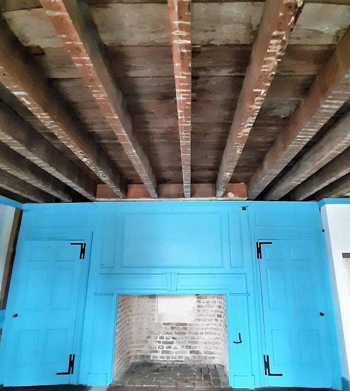 The interior of Hampton shows large rooms with wooden floors. The paint is blue. The fireplace is brick, with chairs sitting around.