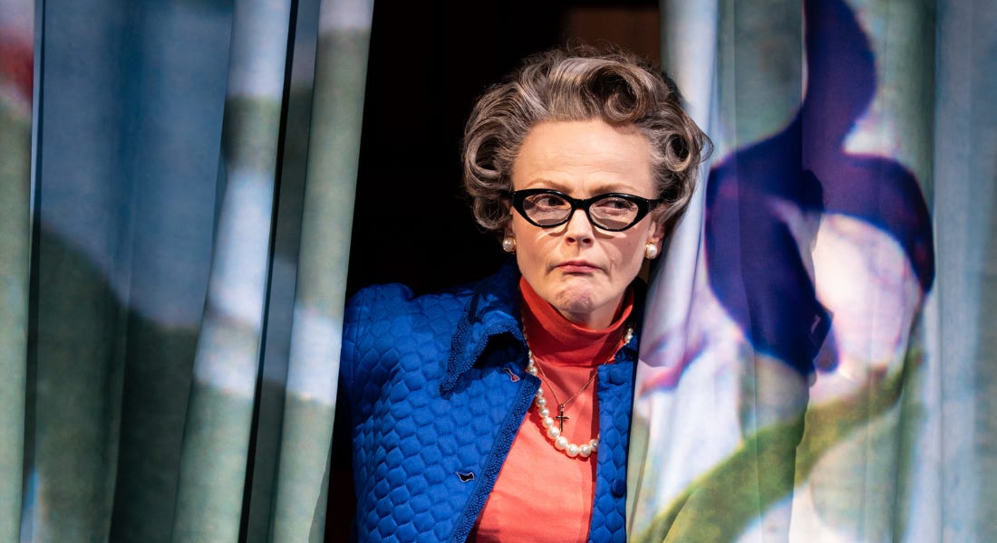 A woman, wearing a blue quilted coat and pink rollneck with dark glasses, stands between two theatre curtains, holding them open. She has a frown on her face and is looking off to the left.