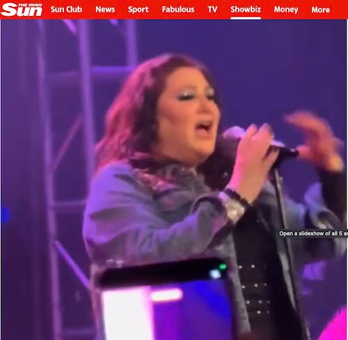 A middle-aged redheaded woman in a denim jacket sings on a stage