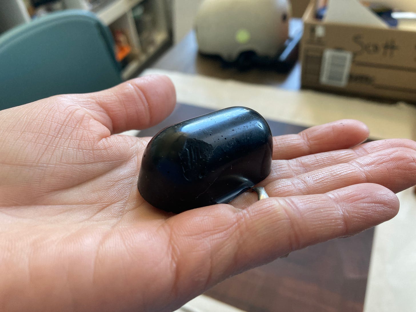 In the palm of a hand, rests a ball of hard ground: A black pill-shaped block that materially seems to resemble a crayon. A copper plate coated in hard ground is in soft focus in the background.
