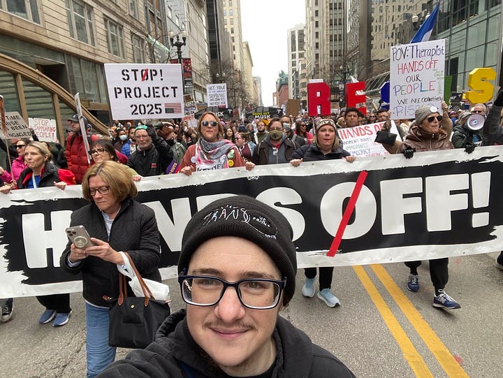 First photo: Asher McMaher (light skin, dark hair, large square glasses, facial hair, black hoodie, black beanie with Trans Up Front logo) marching in protest ahead of long HANDS OFF! banner with large crowd. Second photo: Charlee Friedman (curly hair, large round brown glasses, big smile, facial hair, light skin, wearing a yellow safety vest) ahead of the line of protest marshals and protesters.
