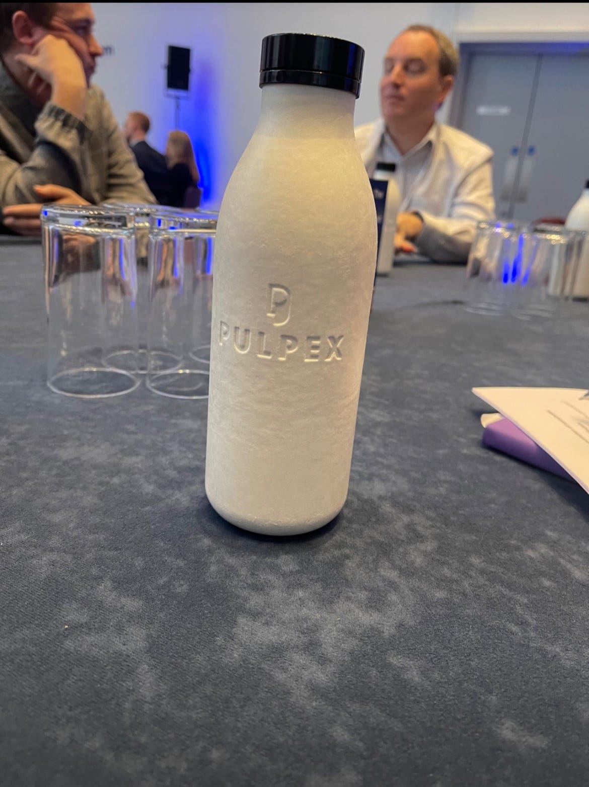 : A close-up of a white, textured bottle with a black cap on a table. The bottle has the logo "PULPEX" embossed on it. In the blurred background, there are people sitting, suggesting the setting is a conference room or a similar indoor event space.