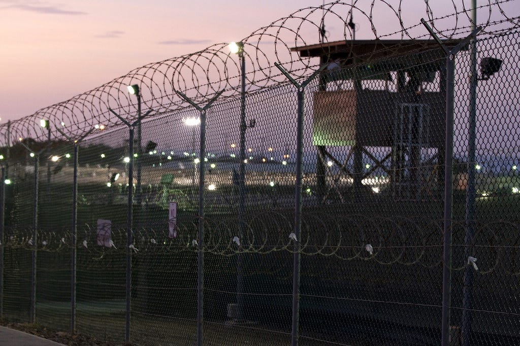 Photo of a guard tower behind a tall fence topped with razor wire, with coils of more concertina wire beyond the fence, silhouetted against a pinkish-purple twilit sky. Pretty artsy for a human rights abuse. Photo of a guard tower behind a tall fence topped with razor wire, with coils of more concertina wire beyond the fence, silhouetted against a pinkish-purple twilit sky. Pretty artsy for a human rights abuse.