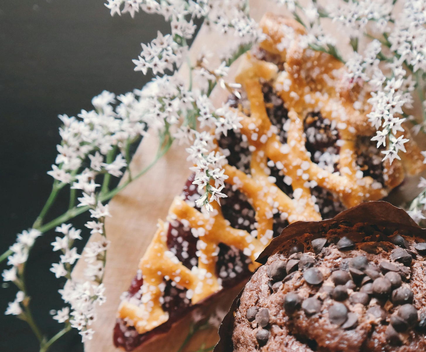 slice of a pie with golden lattice top and dark purple filling on brown paper on dark background covered with blossom branch and next to chocolate muffin slice of a pie with golden lattice top and dark purple filling on brown paper on dark background covered with blossom branch and next to chocolate muffin