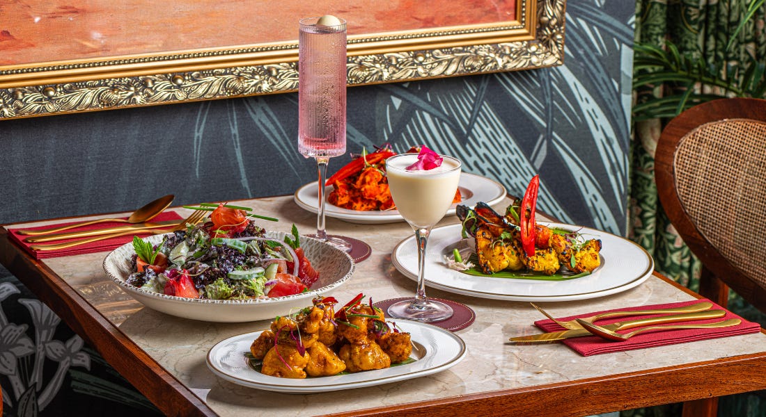 A table with plates of Indian food and salads, two cocktails, and a gold framed painting A table with plates of Indian food and salads, two cocktails, and a gold framed painting