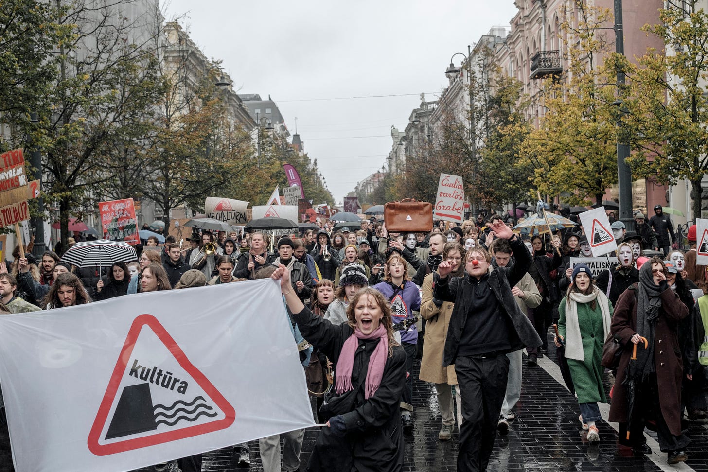 Crowd of people march in the rain in protest. A woman at the front holds a banner that shows a red triangular road sign with the word "kultura" sliding off a cliff into waves