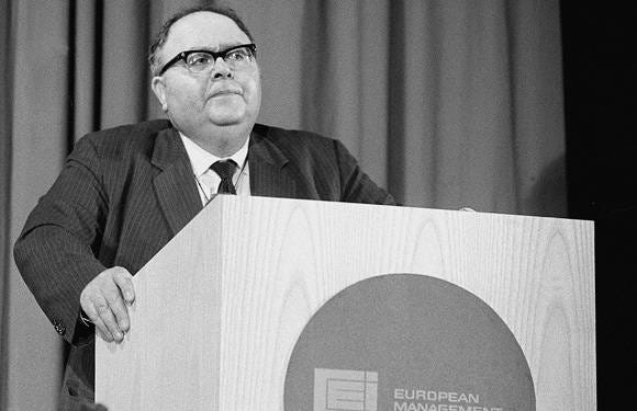 An image of Herman Kahn, a white man, gripping a dais; he has a dark suit and black tie on