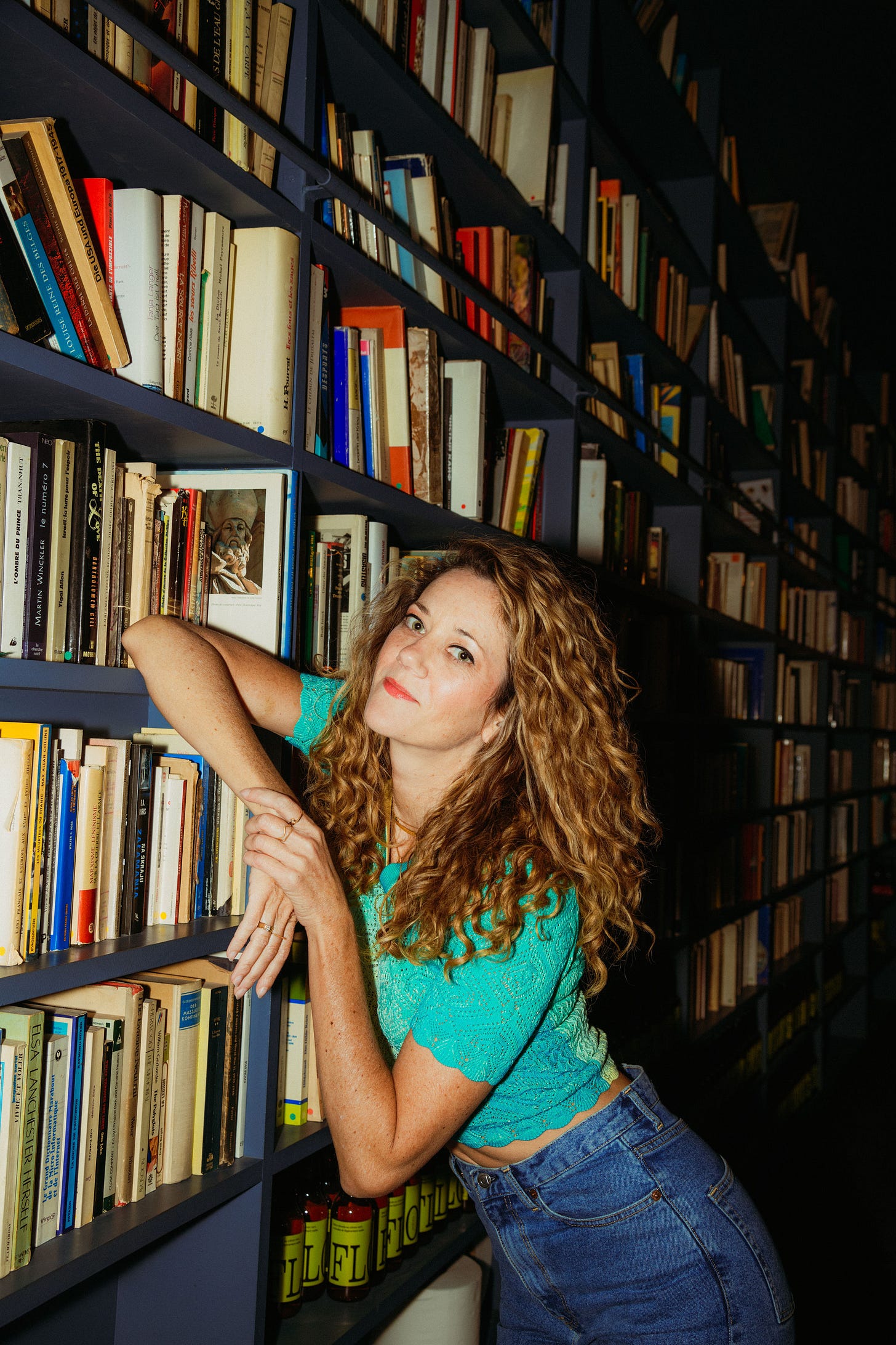 Woman in turquoise shirt with big curly hair leans against a full book case. 
