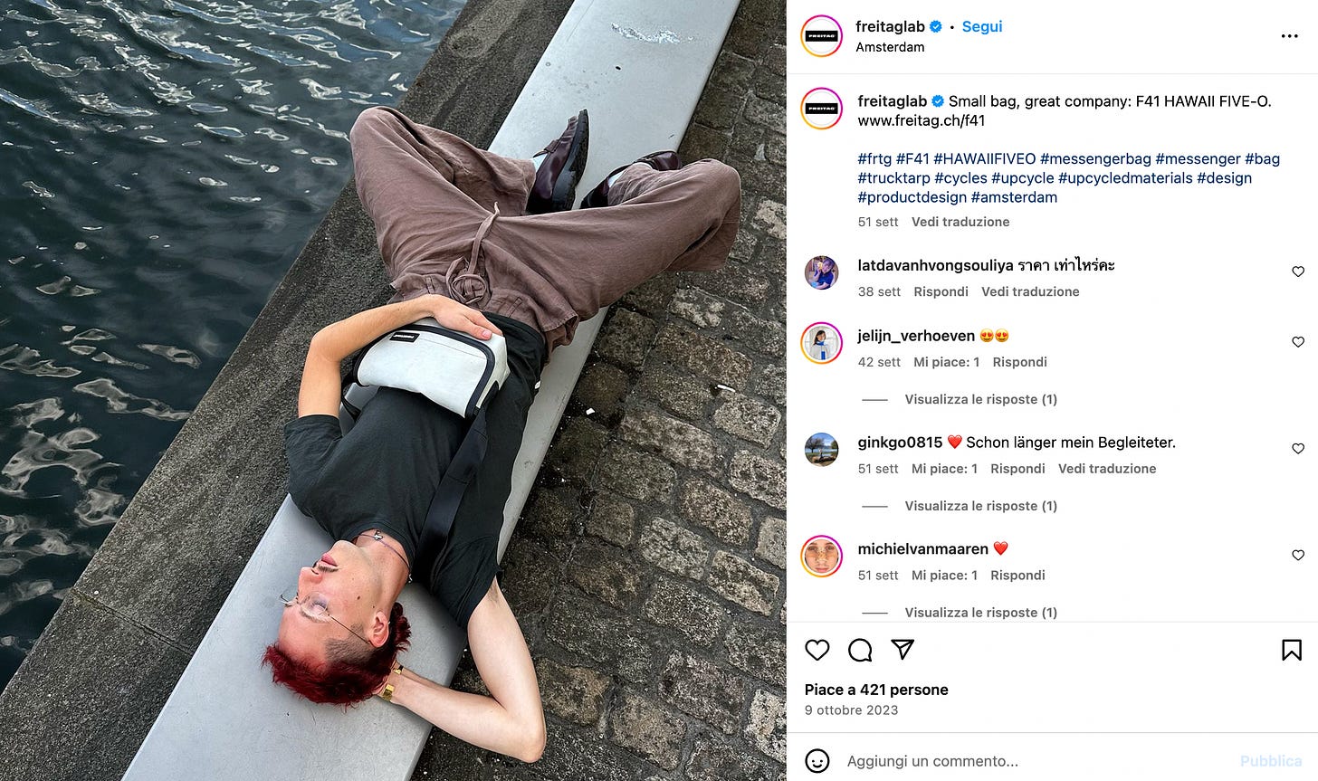 Il soggetto della foto è disteso su un muretto, in città, nei pressi di un fiume. Capelli rossi, muppet, baffetto, occhiali e ciglia lunghissime. Ha una borsa FREITAG poggiata sul ventre. Le gambe a mo' di farfalla, il braccio dietro la nuca e l'altro a sostenere la borsa. Non si capisce se sia uomo o donna, ma questo non ci interessa: si sta rilassando e godendo la sua vita, la sua borsa, la sua libertà di espressione.