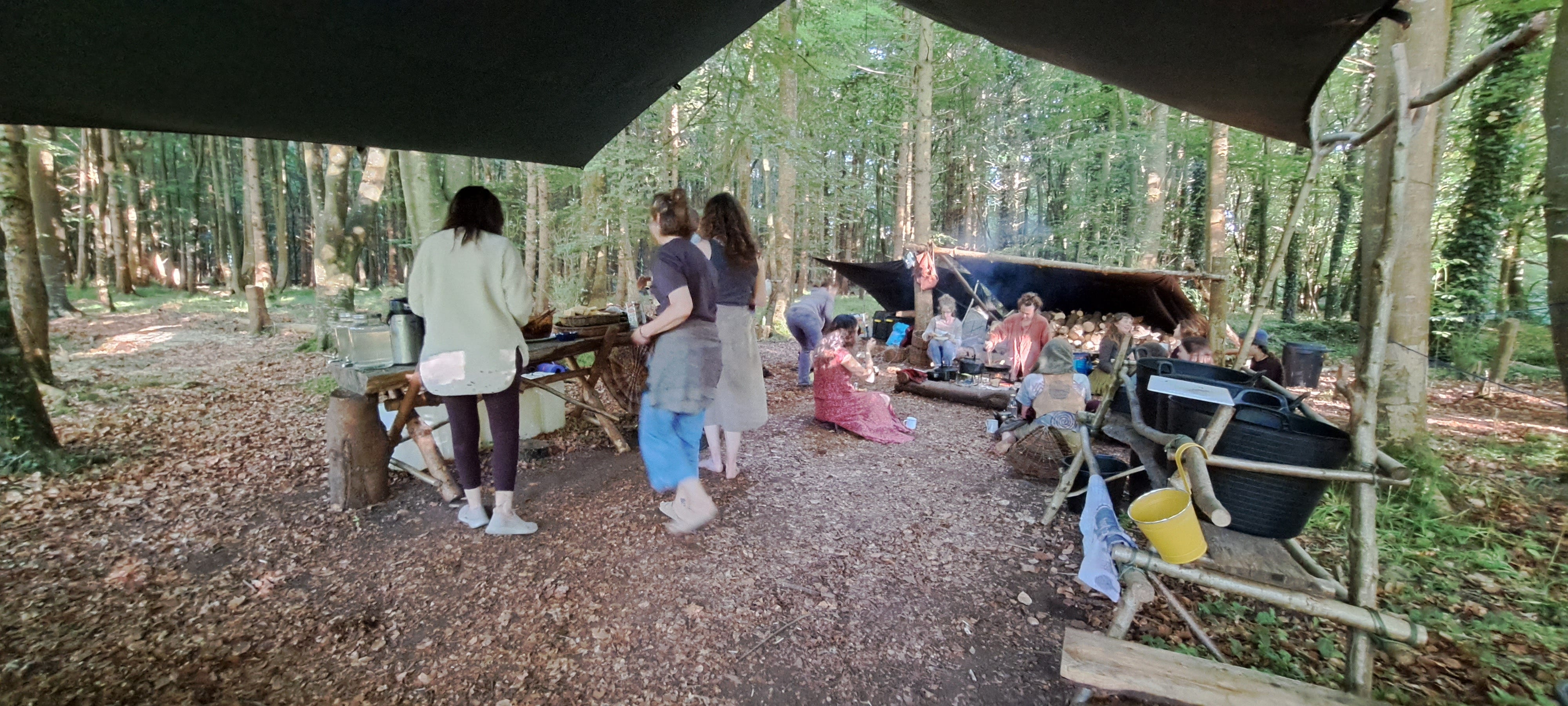 A community gathering space with the hearth in front and the woodpile beyond. To the right is the washing up station and to the left a long trestle table where breakfast is layed out buffet-style. 3 people are standing here, more are seated around the fire. The forest provides a wall of trees and greenery as a backdrop, but the sun is reaching through to spangle the floor with light.