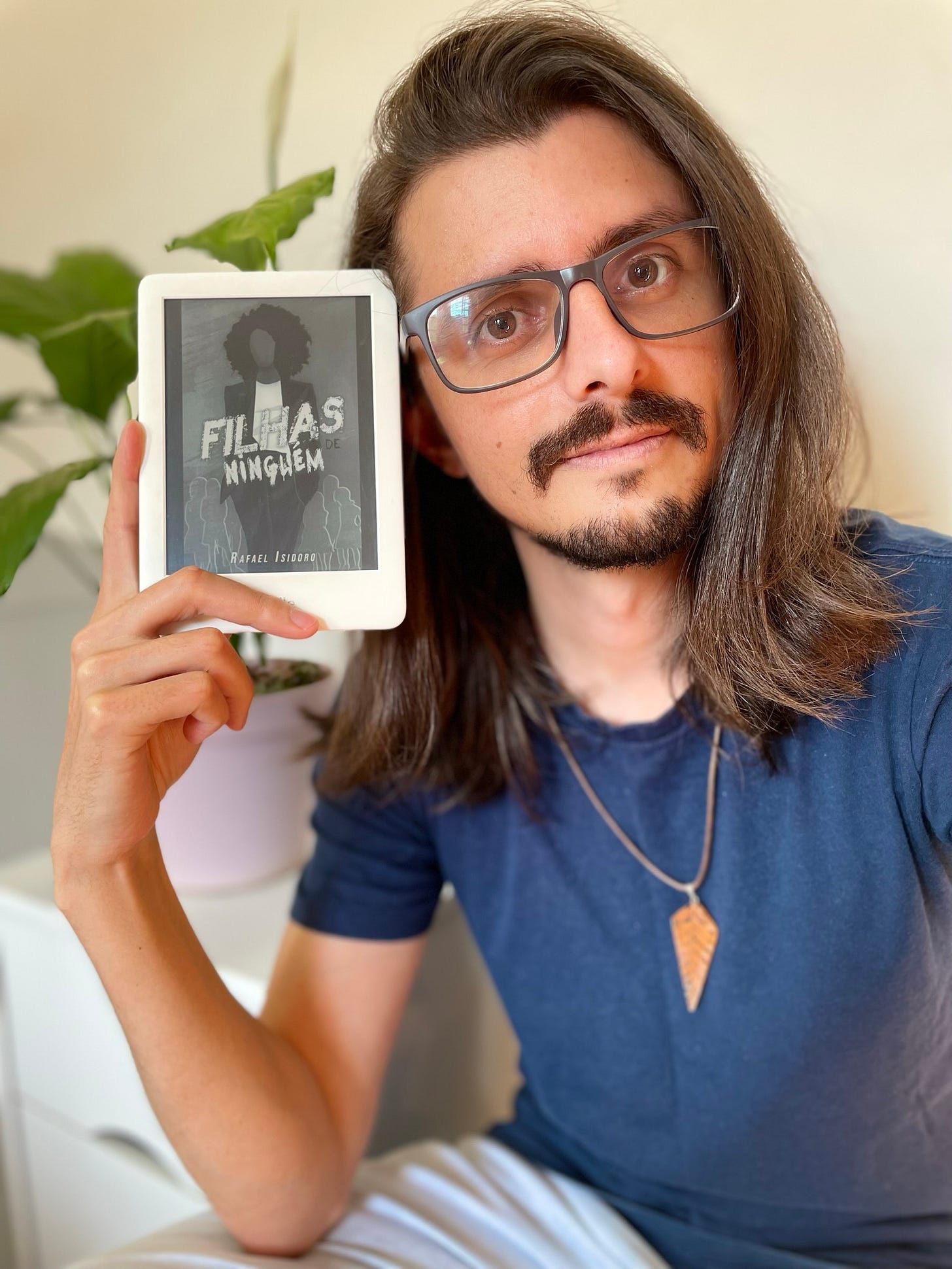 foto do autor de cabelo solto e com o kindle ao lado da cabeça. ele usa uma camisa azul escura e está com o cabelo solto. no kindle, a capa do seu livro "filhas de ninguém"