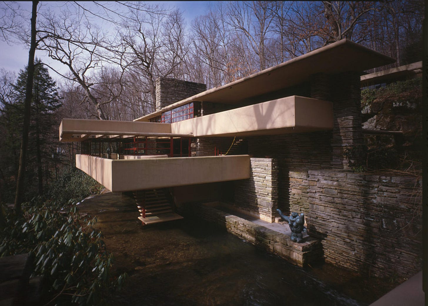 Photo of a modern house with stone walls and cantilevered terraces over a stream surrounded by bare trees.