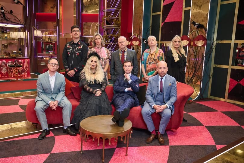 Phil Wang, Lou Sanders, Jack Dee, Harriet Dyer and Daisy May Cooper standing up in front of a couch, with Reece Shearsmith, Roisin Conaty, Jimmy Carr and Tom Allen sitting on it. They are in a large room with a red and black colour pattern and Roisin is wearing a gothic black dress. Phil Wang, Lou Sanders, Jack Dee, Harriet Dyer and Daisy May Cooper standing up in front of a couch, with Reece Shearsmith, Roisin Conaty, Jimmy Carr and Tom Allen sitting on it. They are in a large room with a red and black colour pattern and Roisin is wearing a gothic black dress.