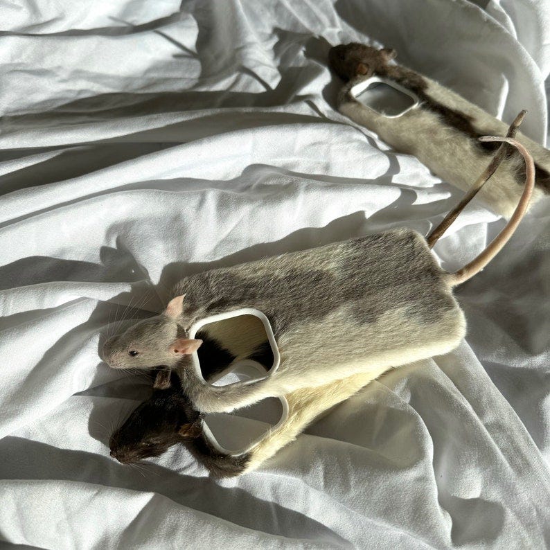 May include: Two gray and white rat-shaped phone cases with white cutouts for the camera lens. The cases are lying on a white fabric surface.