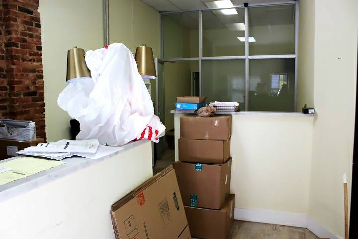 an image of a messy office with two lamps and a plastic bag in the foreground, stacked boxes in the middleground, and a glass office partition in the background