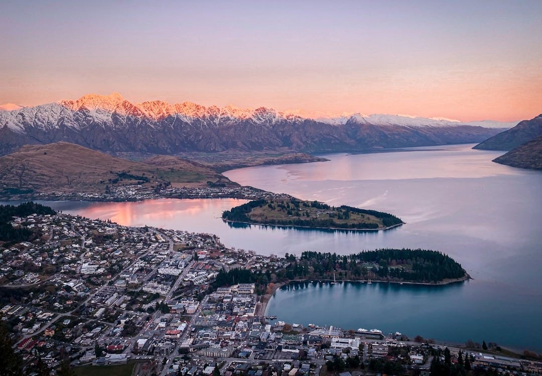aerial view of lake and mountains during daytimeView over Queenstown and Lake Wakatipu at dusk, with the Remarkables mountain range capped in snow behind.