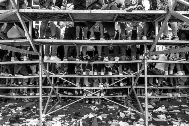 Photographie prise sous la tribune d'un stade improvisé : on ne voit, de dos, que les pieds de dizaines d'enfants assis dans les travées.