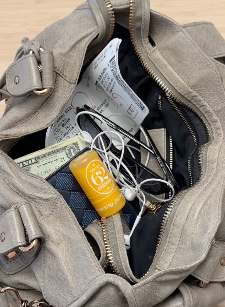 top-down view inside an open, grey leather tote bag. The bag is packed with travel essentials including white wired earphones, US dollar bills, eyeglasses, airline boarding passes, and a small black checkered item. A prominent yellow Sol de Janeiro 'CHEIROSA 62' jelly perfume balm stick is visible near the center.
