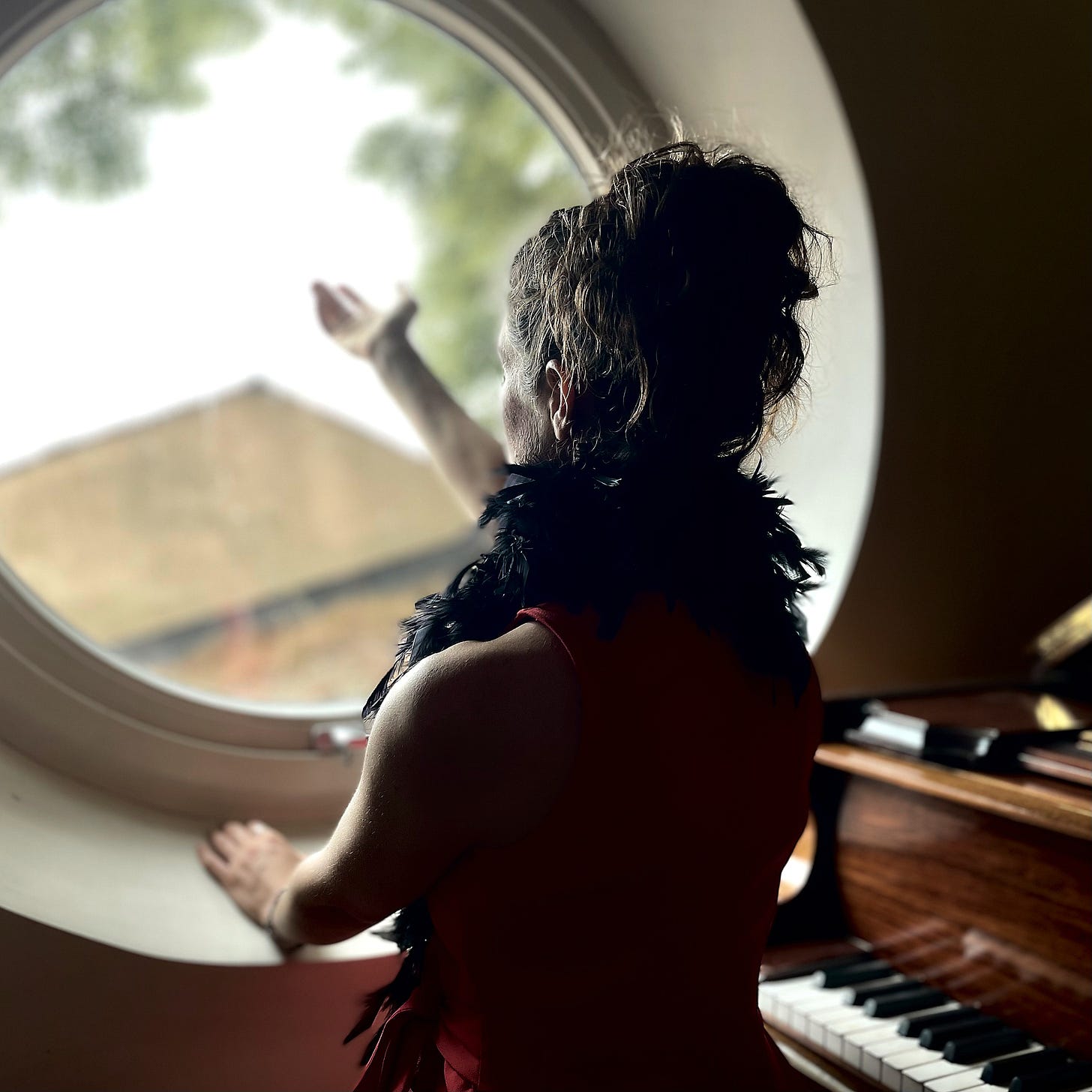 Danielle Styles of Bird Flies Free, standing next to a grand piano and looking out of a circular window, holding up her hand as if to release a bird out of the window. Danielle Styles of Bird Flies Free, standing next to a grand piano and looking out of a circular window, holding up her hand as if to release a bird out of the window.