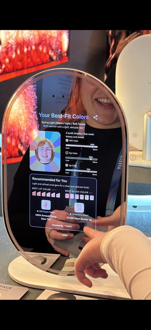 A Samsung AI Mirror displays a digital analysis of a person’s face, showing recommended makeup shades and best-fit colors. Another frame the mirror presents a skin analysis panel with categories like wrinkles, pores, redness, and elasticity. The person’s reflection appears behind the diagnostic overlay while they interact with the device. The reflection of the person smiling is visible behind the transparent interface.