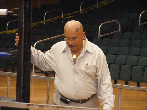 Johnny Rodz, wearing glasses and a light button-down shirt, stands at ringside with one hand on a ring post at a 2008 wrestling show in Albany, New York