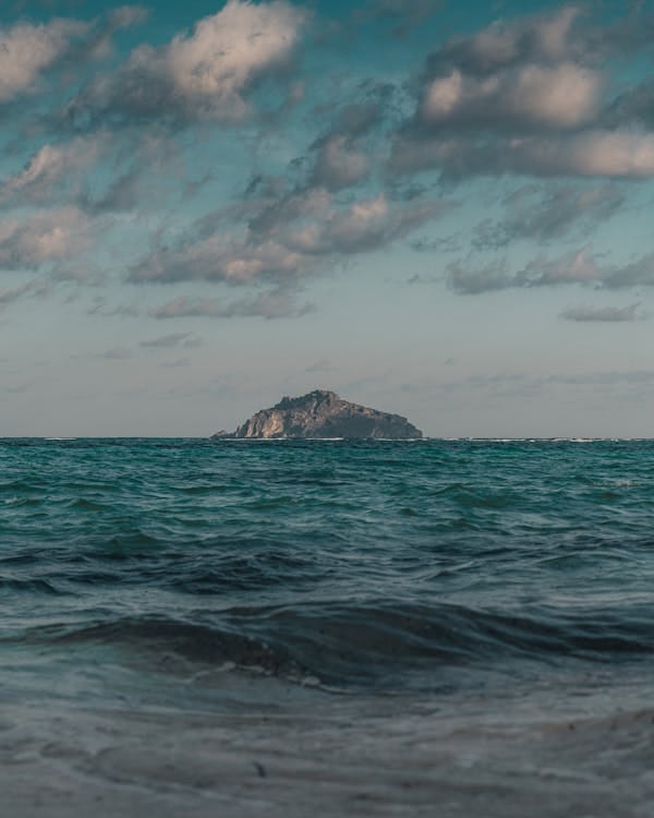 Free A scenic view of a distant island surrounded by vibrant blue ocean under partly cloudy skies. Stock Photo