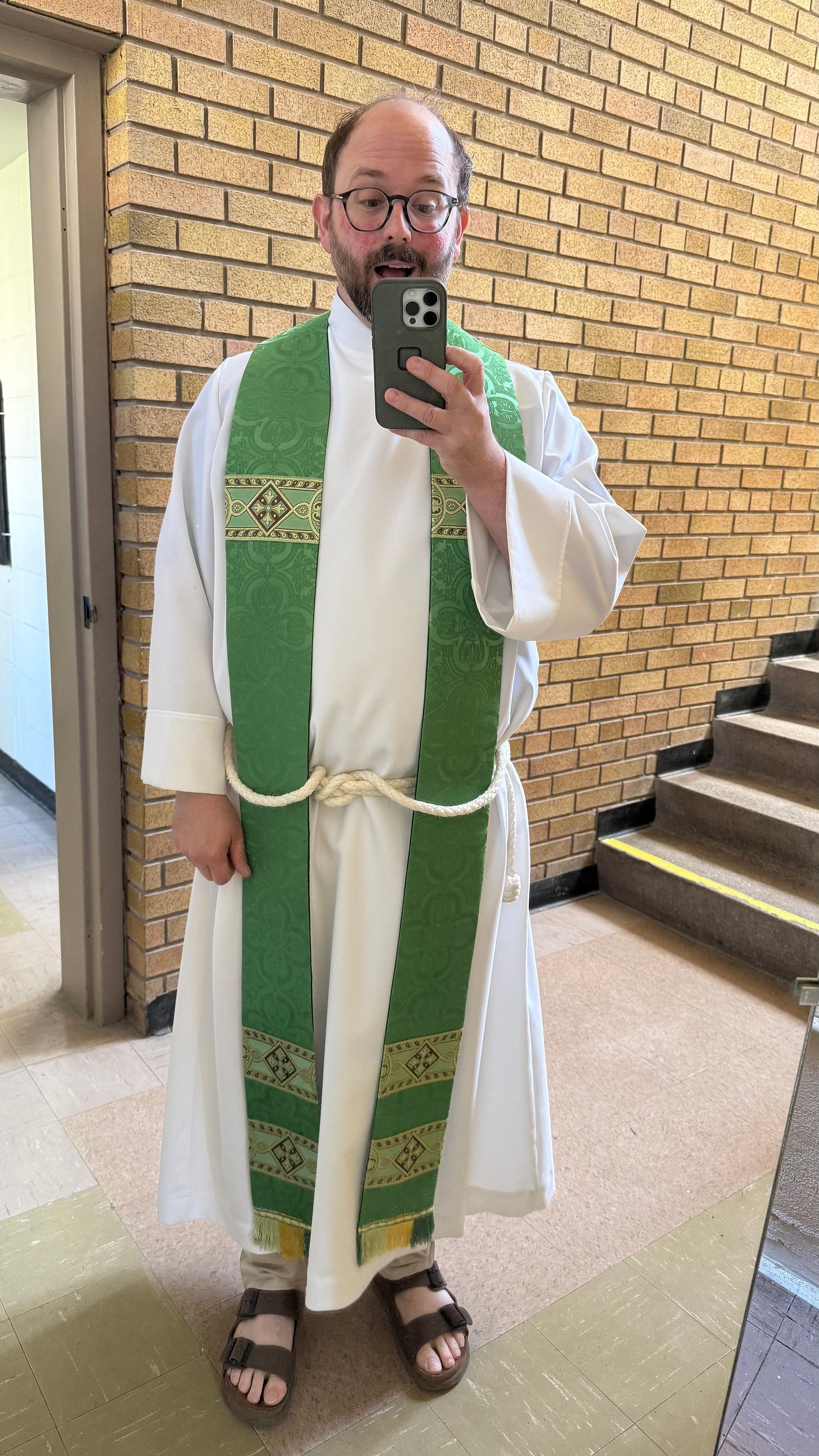 The author goofily taking a selfie while wearing his white cassock-alb and green priest's stole, against a backdrop of brown bricks