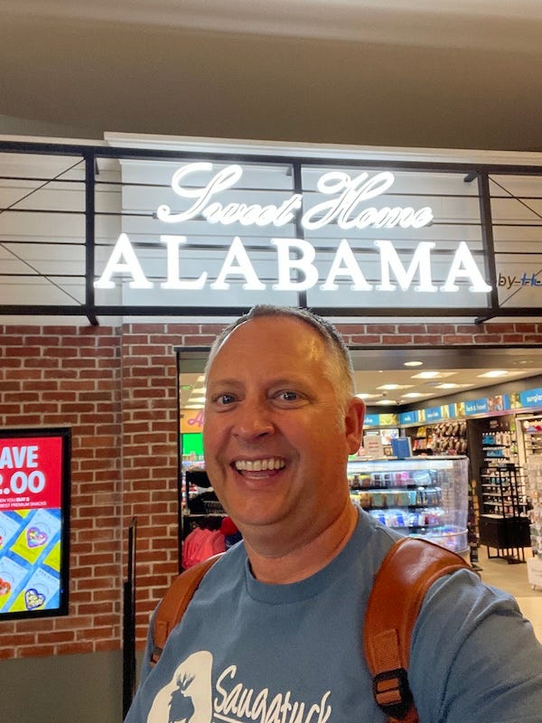 Dr. Dave in front of a sign in the Birmingham airport that says "Sweet Home Alabama"