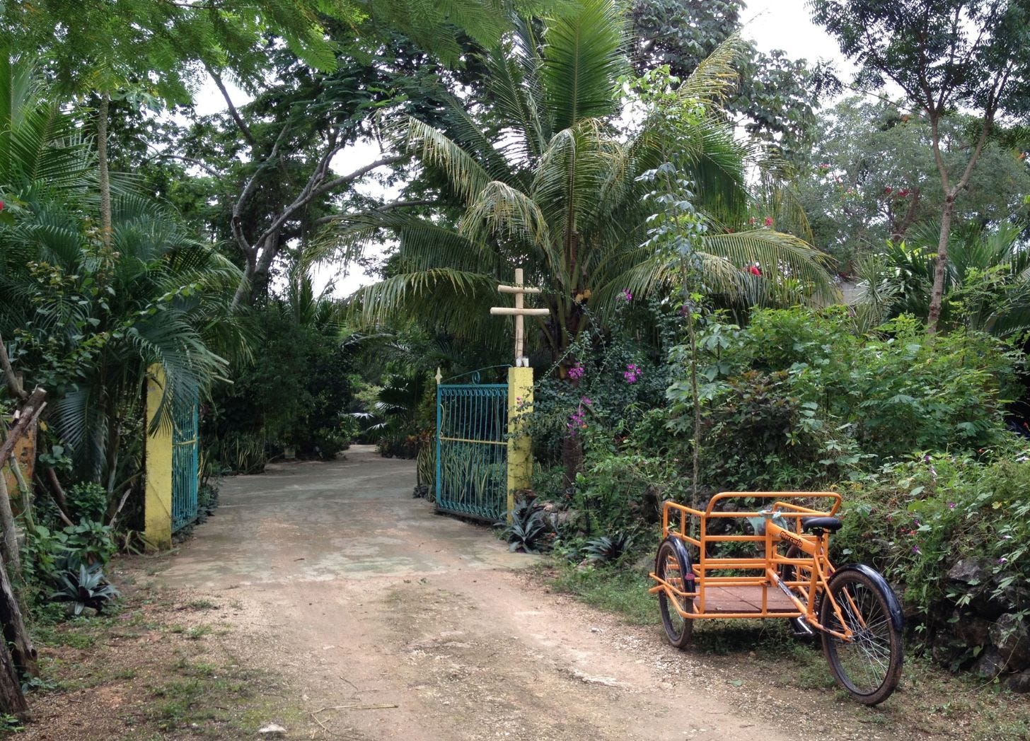 Open iron gate at a rancho in Yucatán, a two-bar wooden cross on a post beside it, an orange cargo tricycle parked on the dirt road. Open iron gate at a rancho in Yucatán, a two-bar wooden cross on a post beside it, an orange cargo tricycle parked on the dirt road.