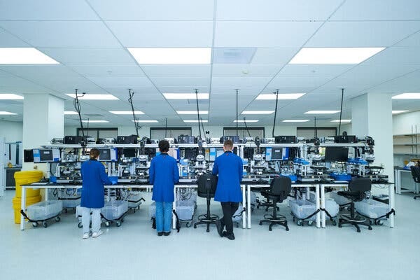 Workers in blue lab coats are seen from the back as they stand at a row of tables packed with lab equipment.