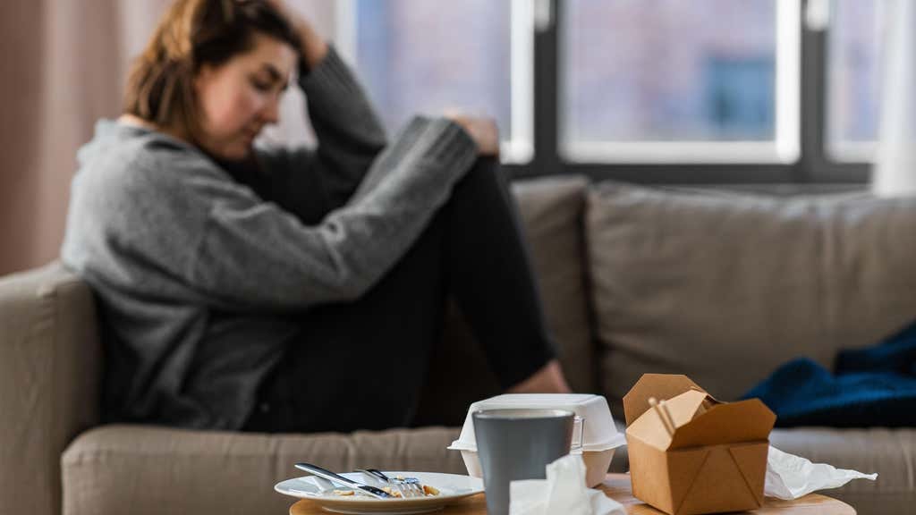 A woman sitting on the sofa, looking unhappy. A plate of food and takeaway cartons are on the table next to her
