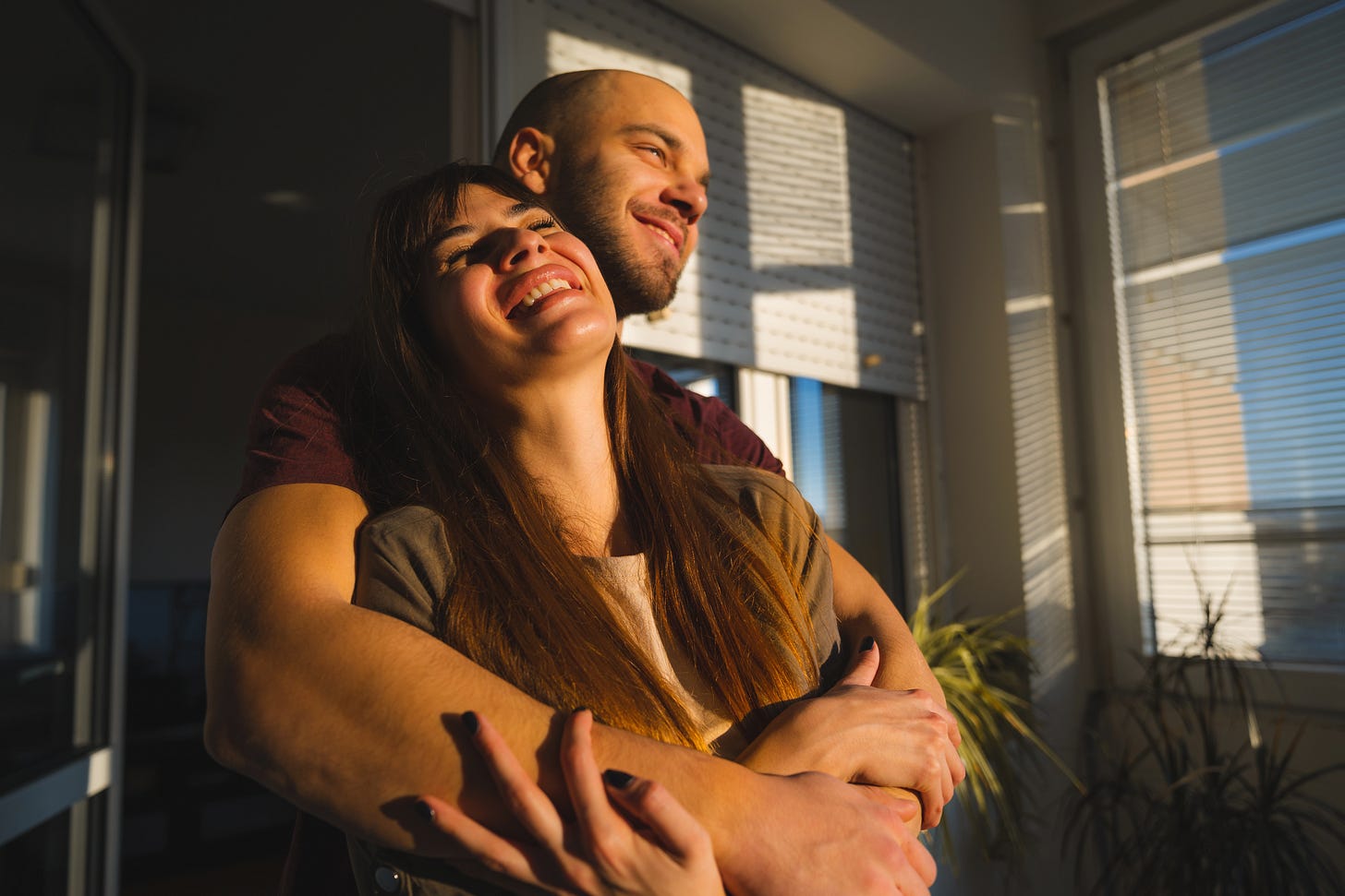 Young man is hugging his girlfriend from behind while she is laughing. Young couple are holding each others in the evening shadows and smiling.
