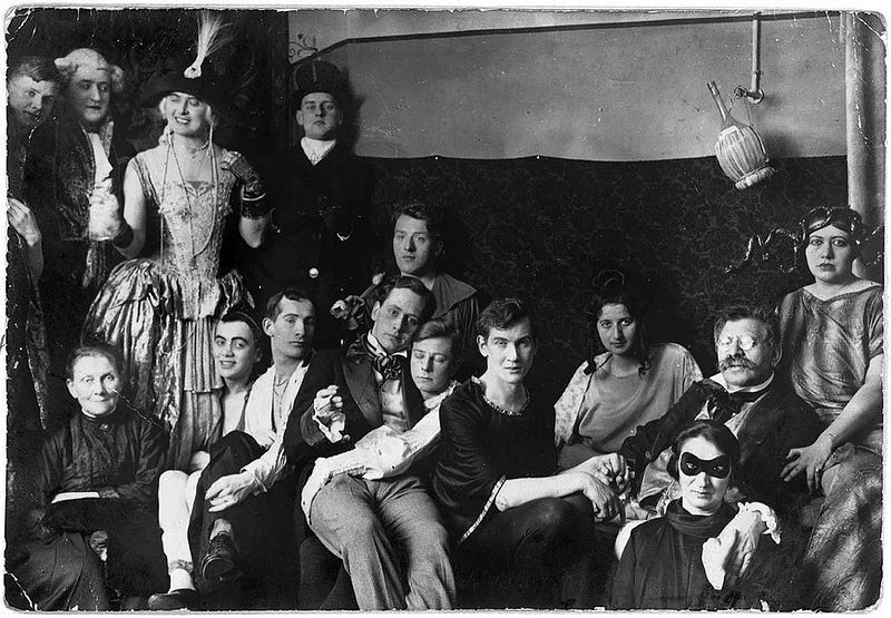 A black and white photo of 15 people dressed in costumes, many hugging or holding hands, looking at the camera. Original caption: Costume party at the Institute for Sexual Research in Berlin, date and photographer unknown. Magnus Hirschfeld (in glasses) holds hands with his partner, Karl Giese (center). Magnus-Hirschfeld-Gesellschaft e.V., Berlin