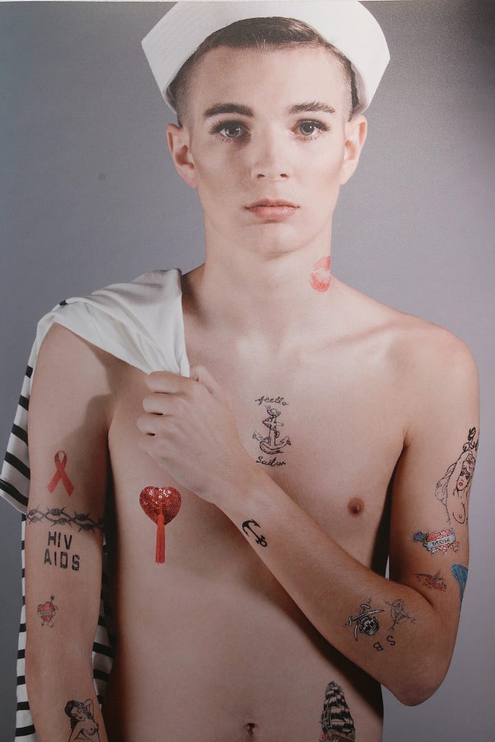 1) Gallery wall with five portraits. The first is a black-and-white image of a two people kissing. It's the final frame of Tessa Boffin's The Sailor and the Showgirl, where they are kissing in front of a Sanyo building. The others show a person in sailor attire and an orange feather boa, exuding confidence. There are 4 modern-day images of the sailor with the boa, the first one, the sailor is shirtless with a heart nipple pastie on. The second image, the sailor is clothed with a striped shirt, and the orange boa. The third image is a full-body image of them holding the sides of their head, and the final image is a mirror image of the sailor holding hands with themself. There is a placard beneath Tessa's work that gives some context about her piece, and the inspired student's piece next to it. 2) Young white skinny person wearing a white sailor hat, holding a white and blue/black striped shirt, with various tattoos including an anchor, ribbon, and barb wire, and HIV AIDS. The expression is neutral. There is a red lipstick print on their neck. They are shirtless, with a red heart nipple pastie over their right nipple. They have short brown hair, and they are staring straight into the camera. They are standing in front of a grey backdrop. They are holding the shirt over their right shoulder.