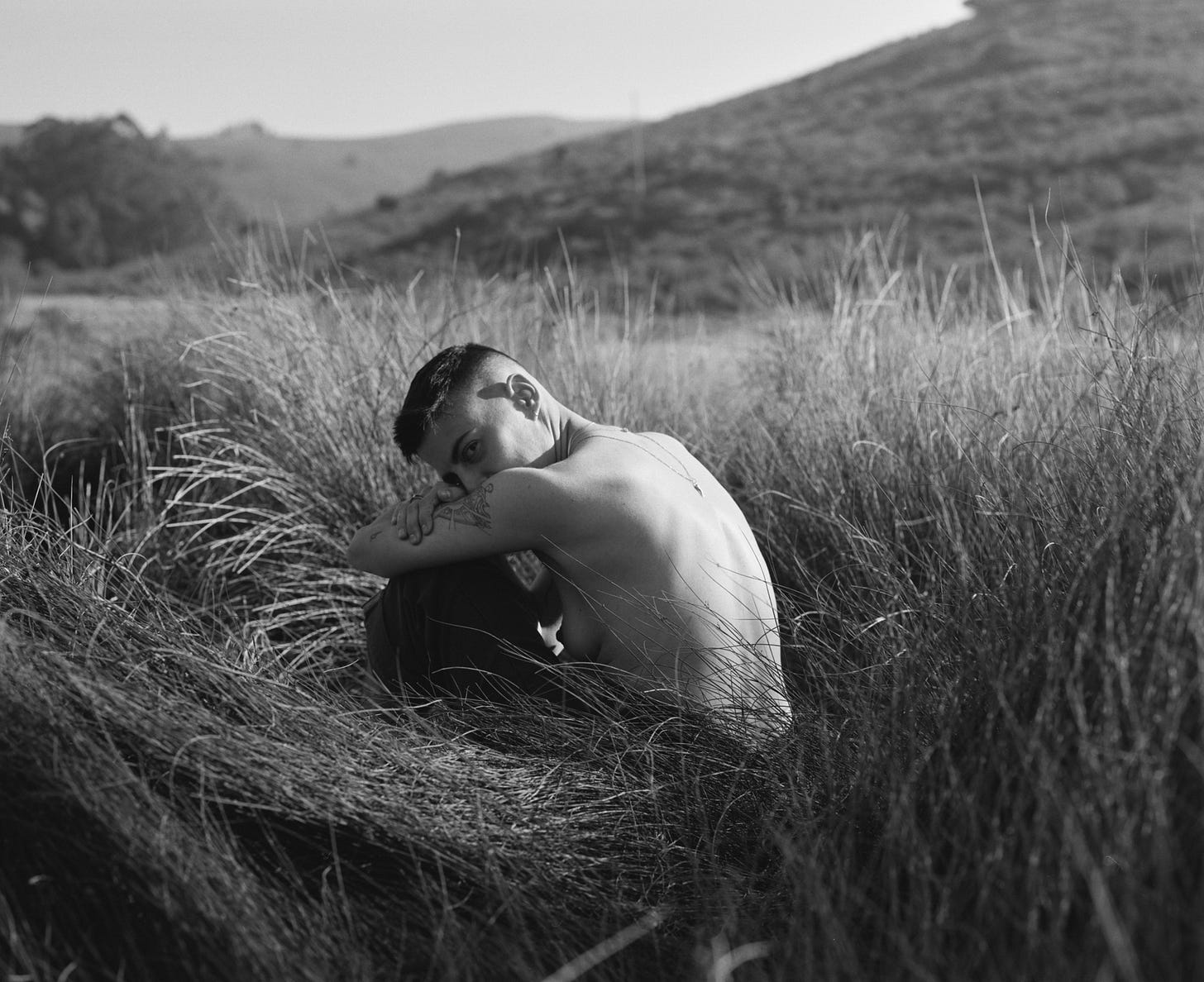A shirtless person with short hair sits curled up in tall grass, resting their head on their arm while looking back over their shoulder. A shirtless person with short hair sits curled up in tall grass, resting their head on their arm while looking back over their shoulder.