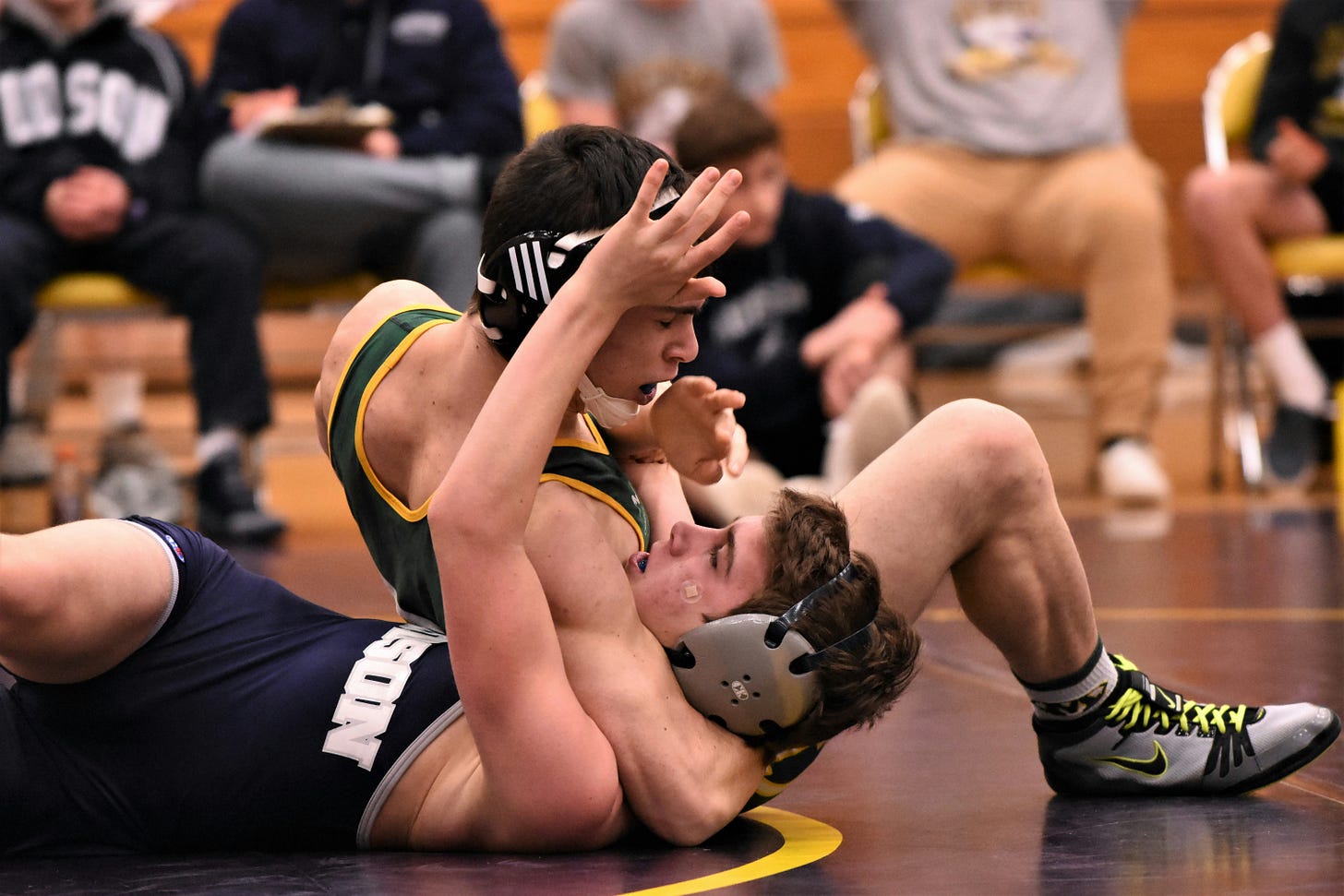 Two wrestlers compete on a dark blue mat with yellow lines; one wrestler is wearing a green and yellow singlet, and he's got a wrestler in a dark blue and gray singlet in a headlock. Two wrestlers compete on a dark blue mat with yellow lines; one wrestler is wearing a green and yellow singlet, and he's got a wrestler in a dark blue and gray singlet in a headlock.