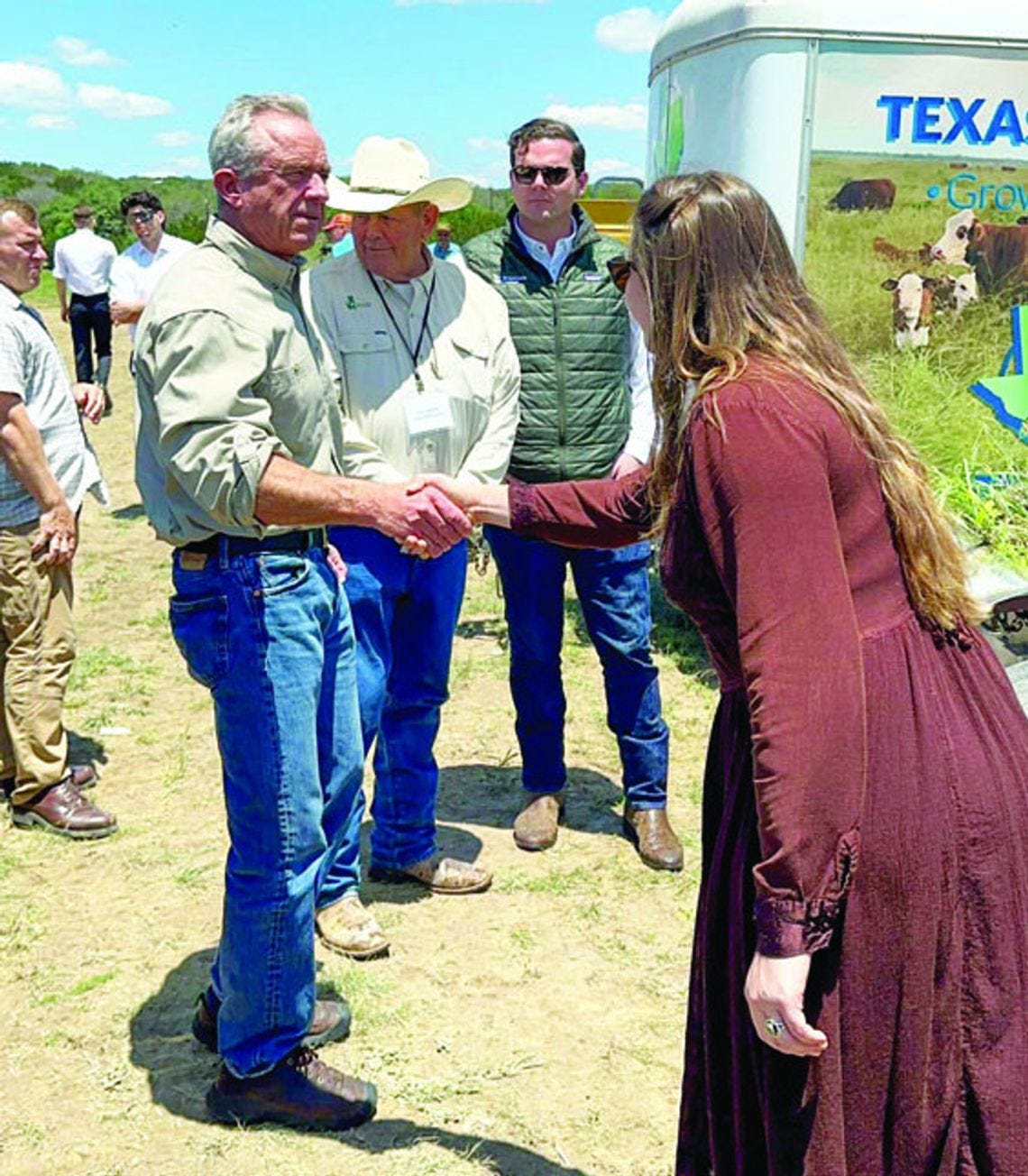  Robert F. Kennedy Jr. shakes hands with a woman in a maroon dress at Sovereignty Ranch with cattle visible behind them.