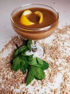 finished malt tonic product. Features brown malt tonic in coup glass with twist of lemon on top. glass is on a white background with malt sprinked around and mint at the bottom of the stem of the glass