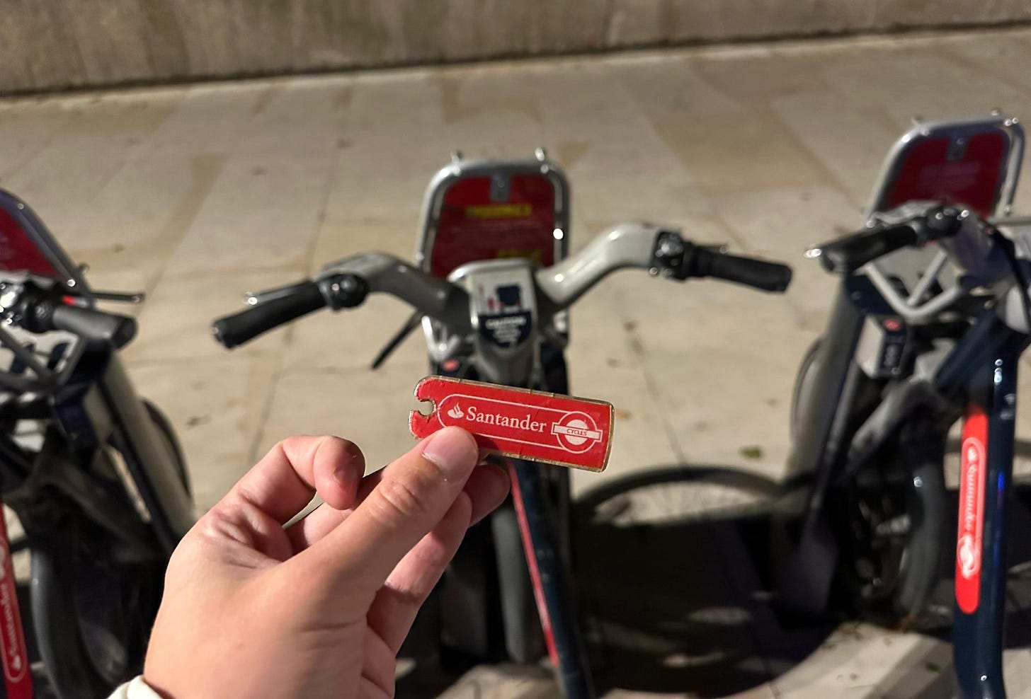 A small red but heavily worn Santander Cycles key is held in the foreground, with docked bikes in the background.
